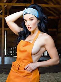 A tousled haired apron clad woman in 1950s style stands in a dusty barn at high noon
